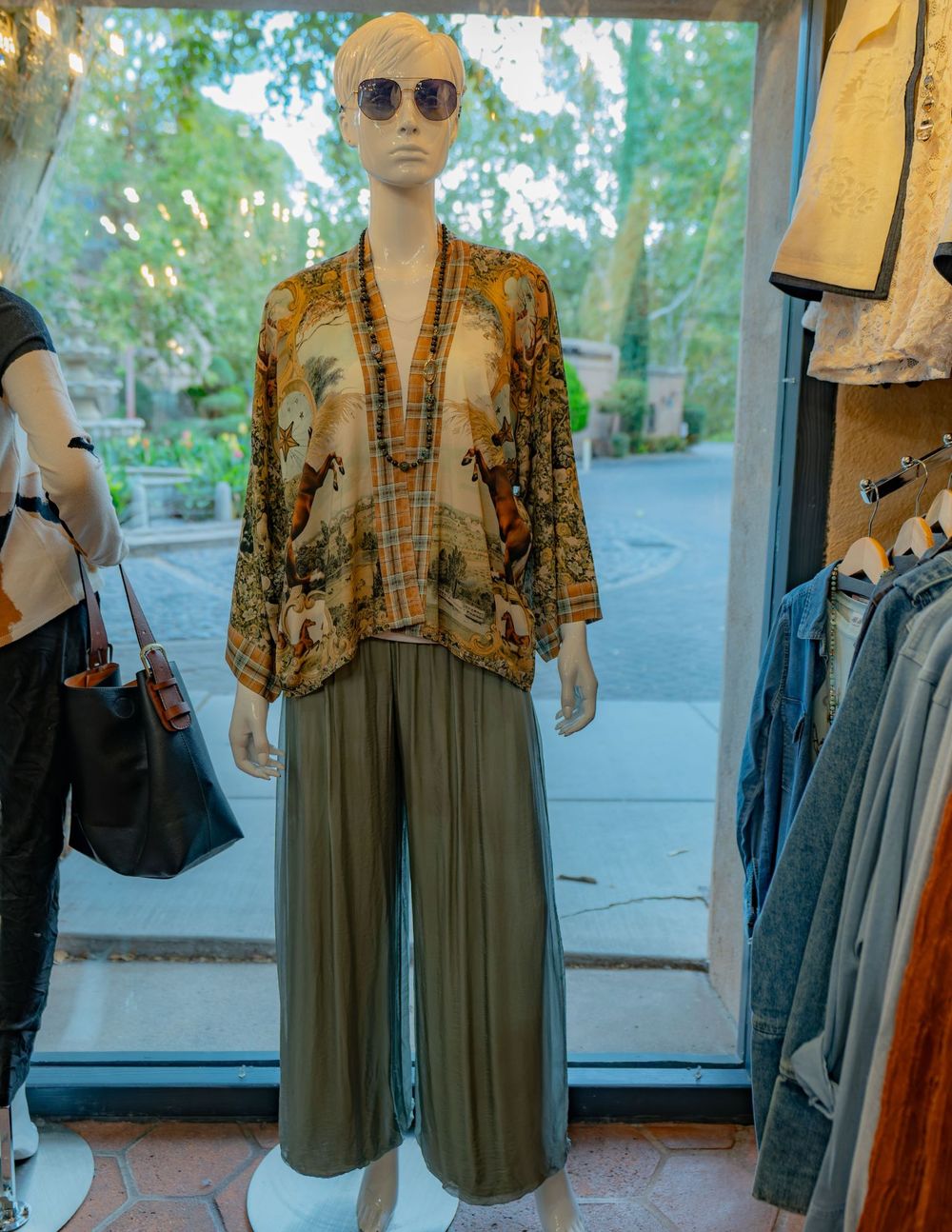 A mannequin in a horse-print kimono jacket and flowing olive silk pants, with the courtyard behind the window