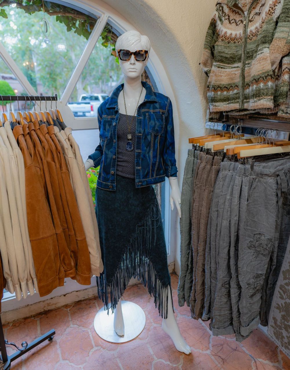 A mannequin in a patterned denim jacket and a long fringed skirt, beside a rack of faux suede jackets