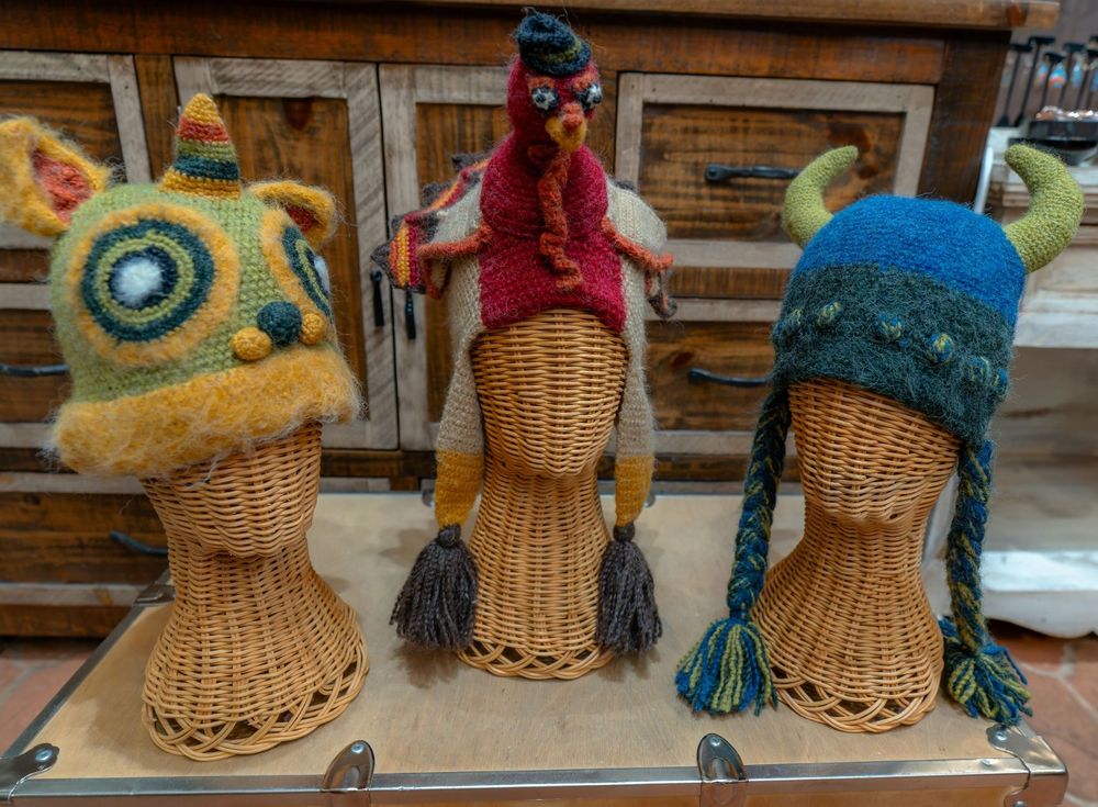 Three hand-knit hats on wicker heads: a green monster with a horn, a red turkey, and a blue monster with horns